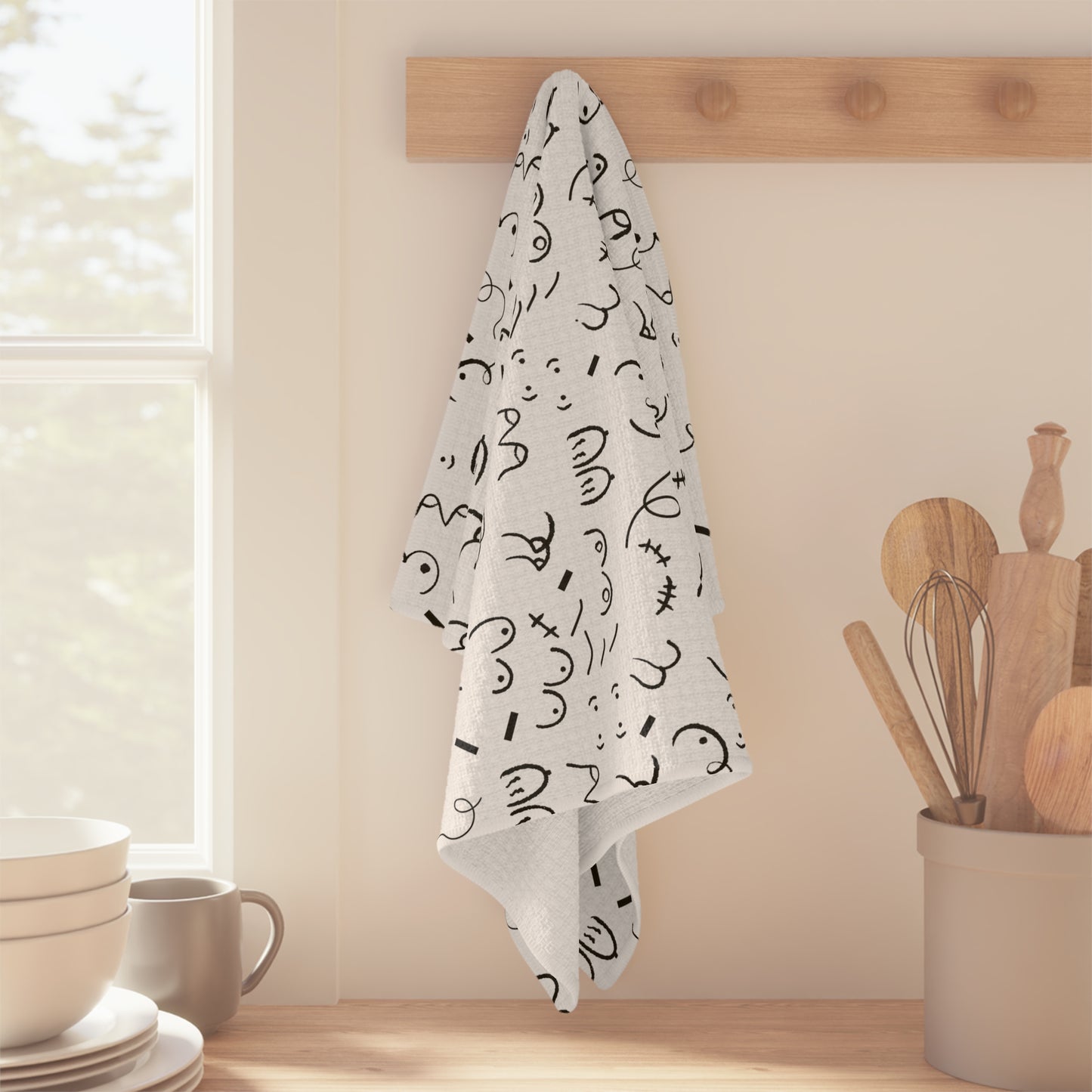 Boobie Pattern Breastfeeding Towel with black abstract designs hanging on a wooden peg rack above a counter with wooden utensils.