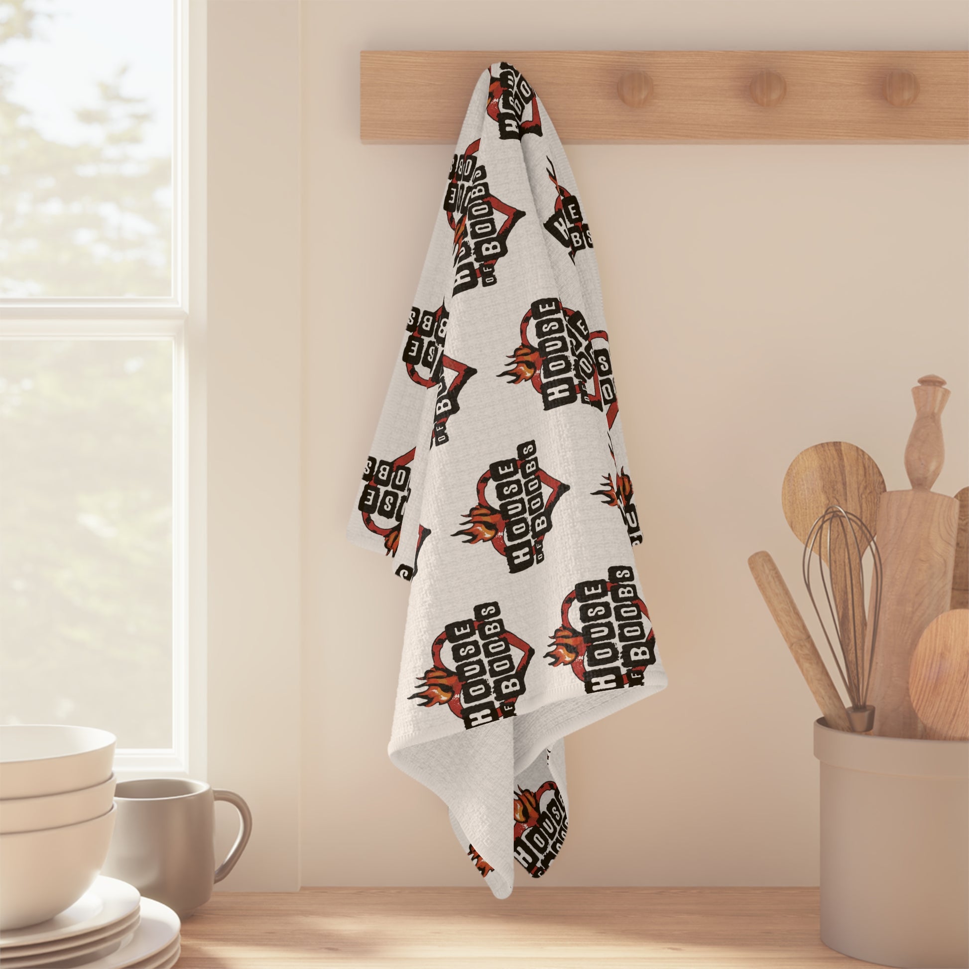 A "House of Boobs" Pattern Breastfeeding Towel, featuring the "HOUSE OF BBQS" logo and a boobie design, hangs on a wooden peg in a kitchen, with utensils and a window in the background.