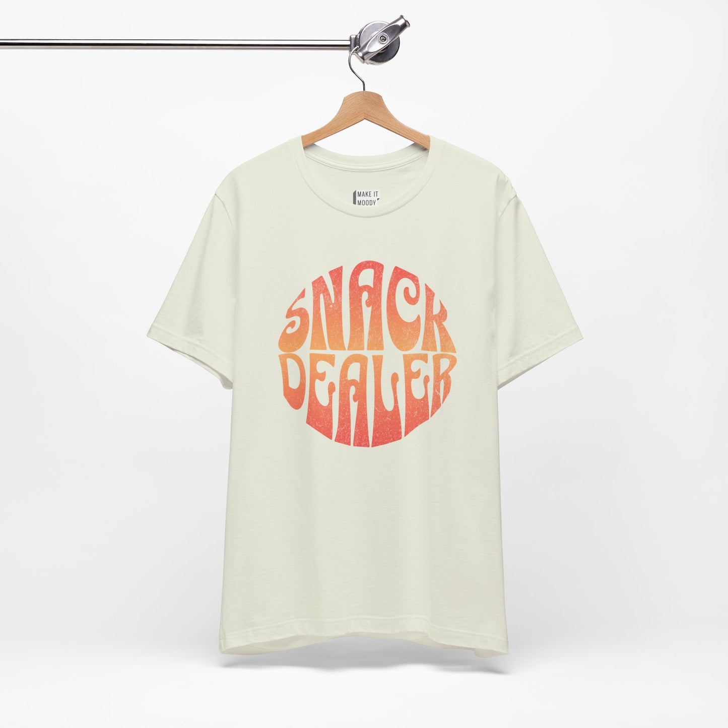 A light green mom t-shirt featuring a retro, orange gradient font with the words SNACK DEALER printed inside a circular design.