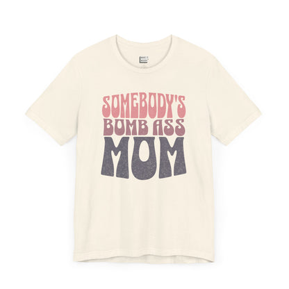 Off-white mom t-shirt featuring bold, retro-style text reading Somebody's Bomb Ass Mom in pink and grey.