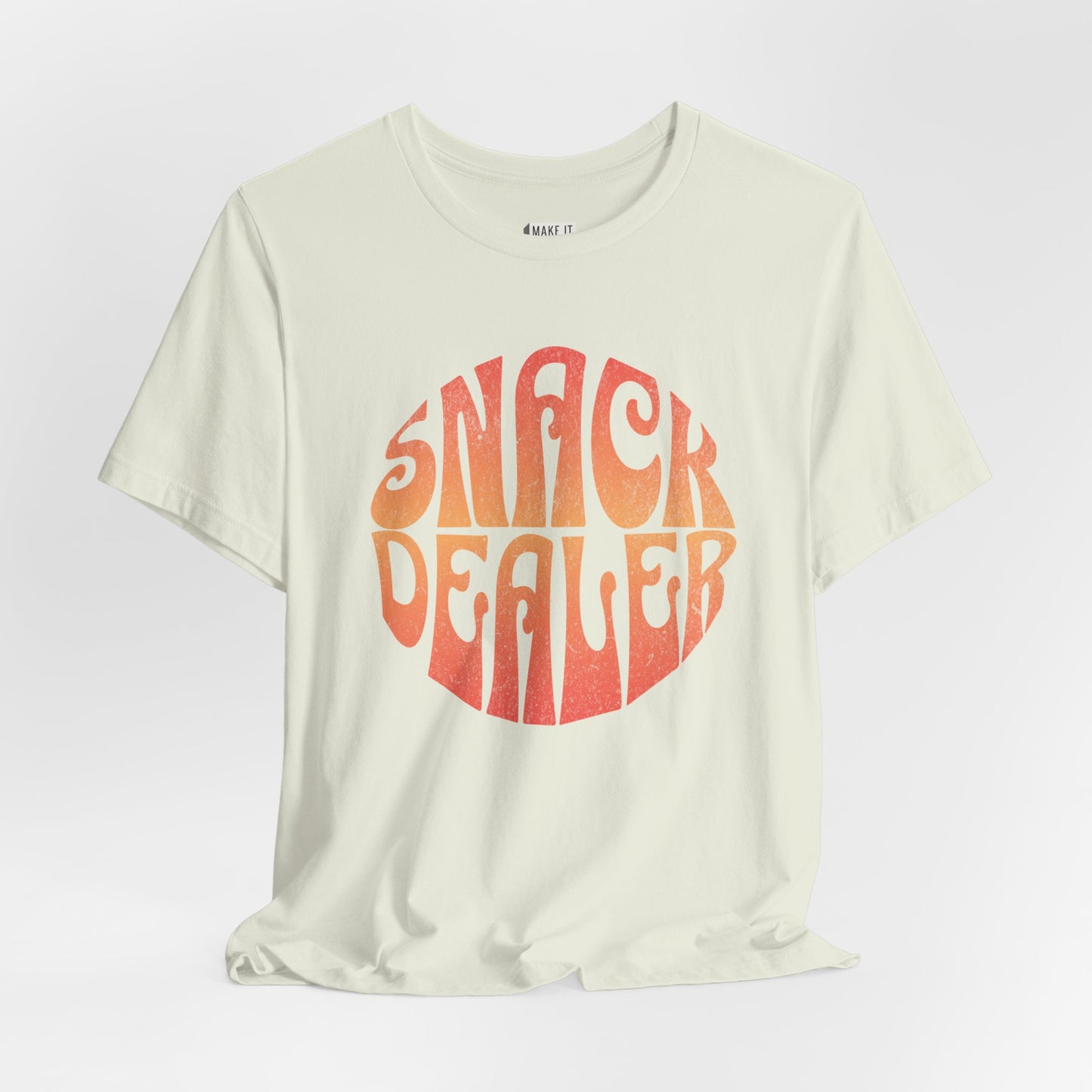 A light green mom t-shirt featuring a retro, orange gradient font with the words SNACK DEALER printed inside a circular design.