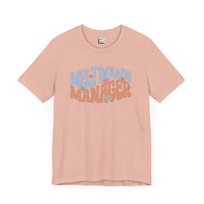 Peach colored mom t-shirt featuring the words MELTDOWN MANAGER in a colorful retro-style font with retro flower designs.