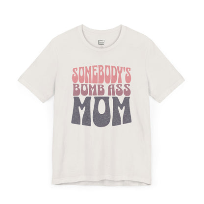 Vintage white mom t-shirt featuring bold, retro-style text reading Somebody's Bomb Ass Mom in pink and grey.