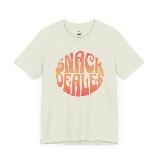 A light green mom t-shirt featuring a retro, orange gradient font with the words SNACK DEALER printed inside a circular design.