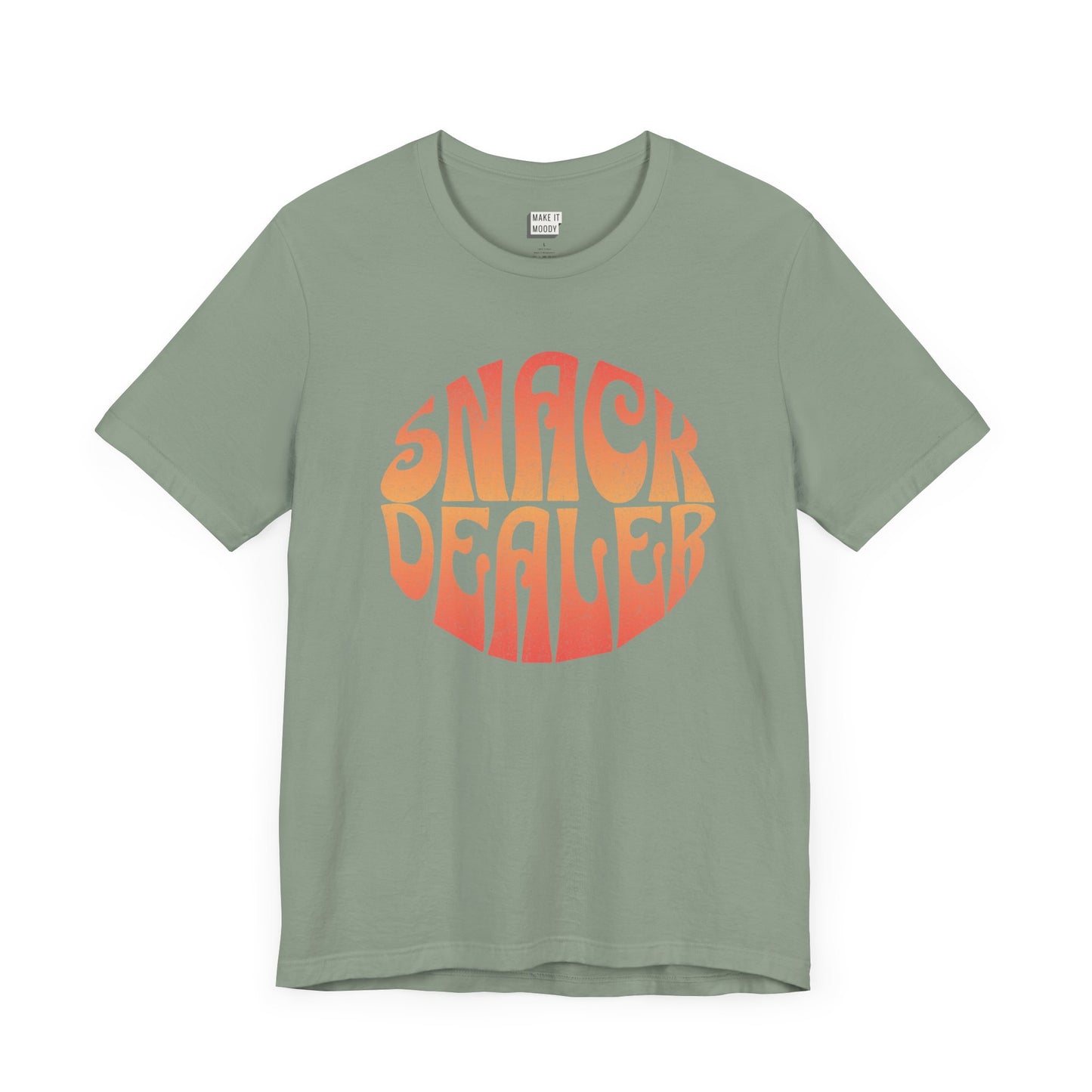 A sage green mom t-shirt featuring a retro, orange gradient font with the words SNACK DEALER printed inside a circular design.