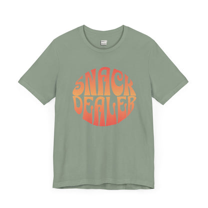 A sage green mom t-shirt featuring a retro, orange gradient font with the words SNACK DEALER printed inside a circular design.