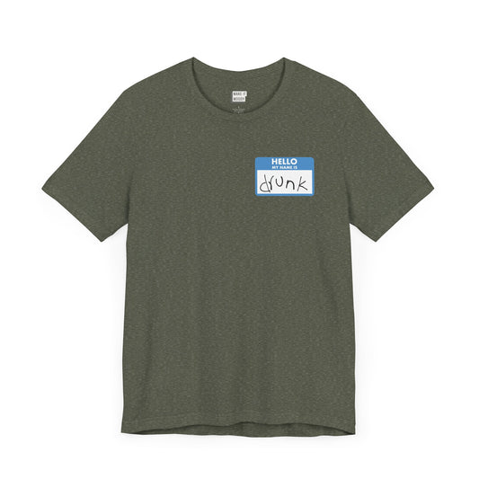 heather military green funny drinking t-shirt with a graphic the resemble and blue sticker name tag on the front in the top left corner. The name tag says HELLO MY NAME IS DRUNK, with the word drunk being handwritten in messy writing.