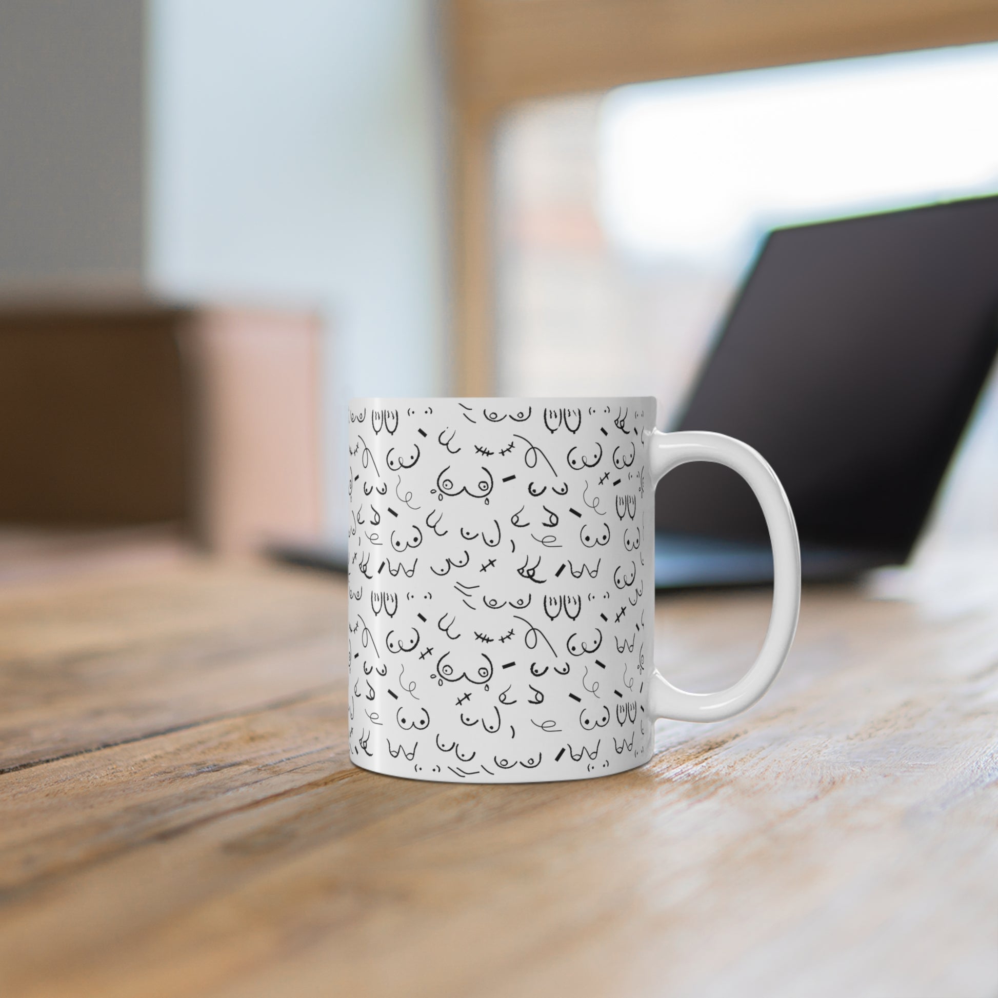 The Boobie Pattern Breastfeeding Mug 11oz, featuring various black doodles including a playful boob pattern, sits on a wooden desk near a black laptop.