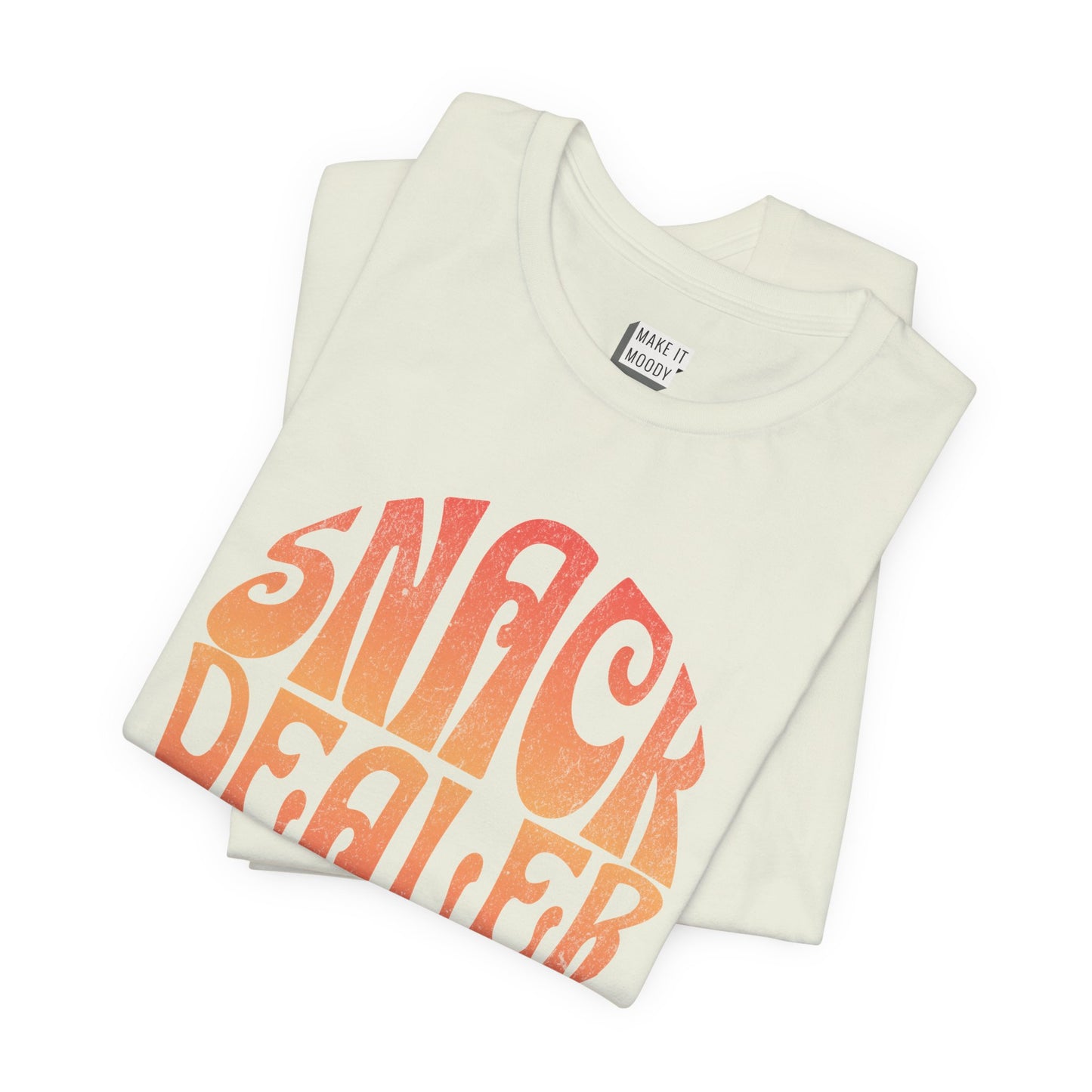 A light green mom t-shirt featuring a retro, orange gradient font with the words SNACK DEALER printed inside a circular design.