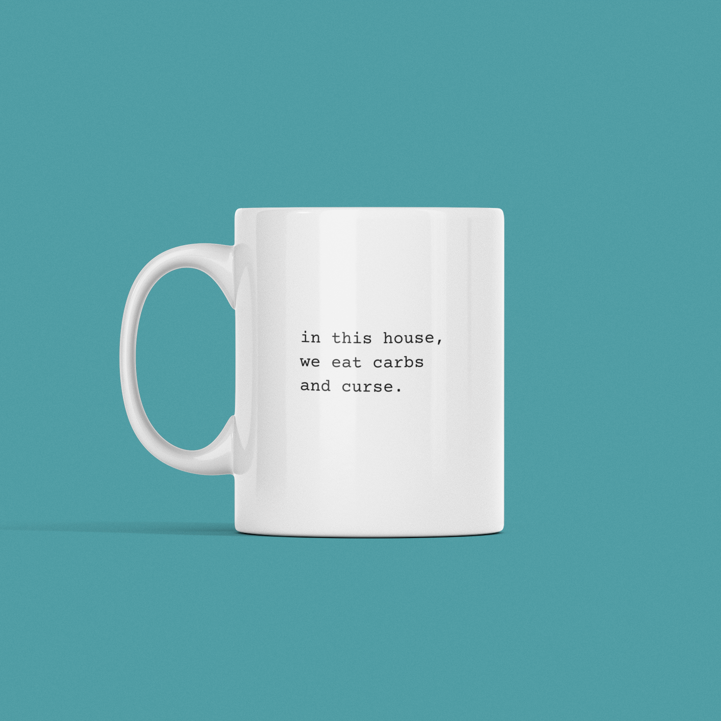 A "In This House" mug on a teal background, featuring the text "in this house, we eat carbs and curse.